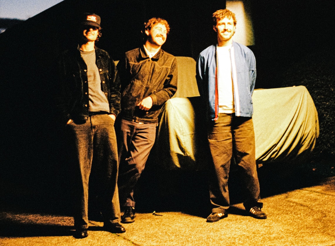 Australian surf rock trio The Grogans standing and smiling outdoors under warm night lighting in a photo by Dylan Jardine.